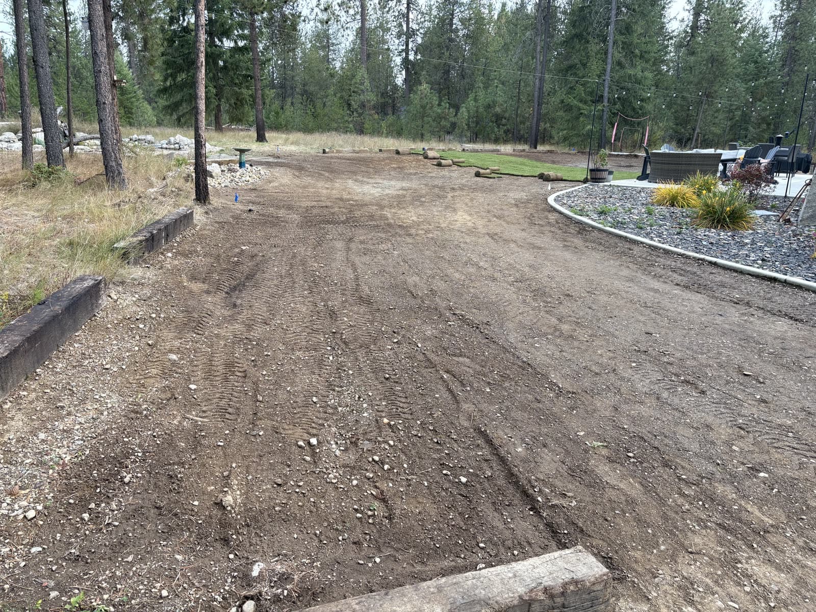 Before: A wooded North Idaho backyard going from bare, graded soil to a finished green sod lawn around a string-lit patio.